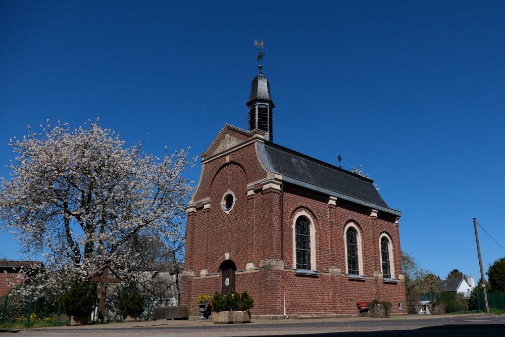 Eine Kapelle aus den für die Region typischen rötlichen Klinkern. An der Längsseite sind 3 oben abgerundete Fenster zu sehen, links an der kürzeren Fassade die Eingangstür. Links neben der Kapelle steht ein großer blühender Baum, im Hintergrund strahlend blauer Himmel.