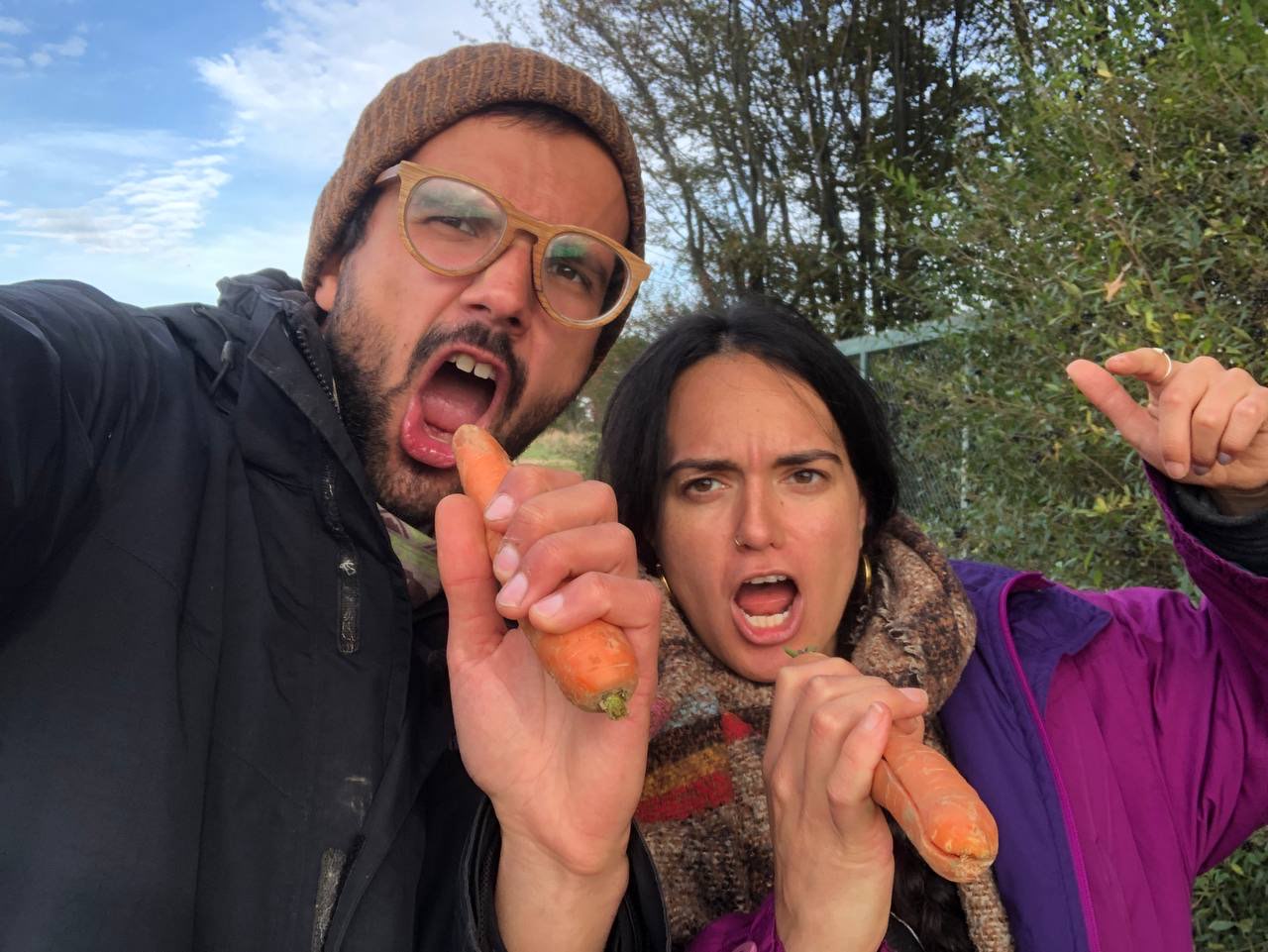 2 Personen, die beide eine Möhre vor den Mund halten und den Mund geöffnet haben, so als würden sie in ein Mikrofon singen. Im Hintergrund sind blauer Himmel sowie Bäume und Gestrüpp zu sehen. Die linke Person trägt eine Mütze, eine Holzbrille und hat einen kurzen Bart. Die Person rechts ist kleiner, hat lange schwarze Haare und trägt einen dicken Schal.