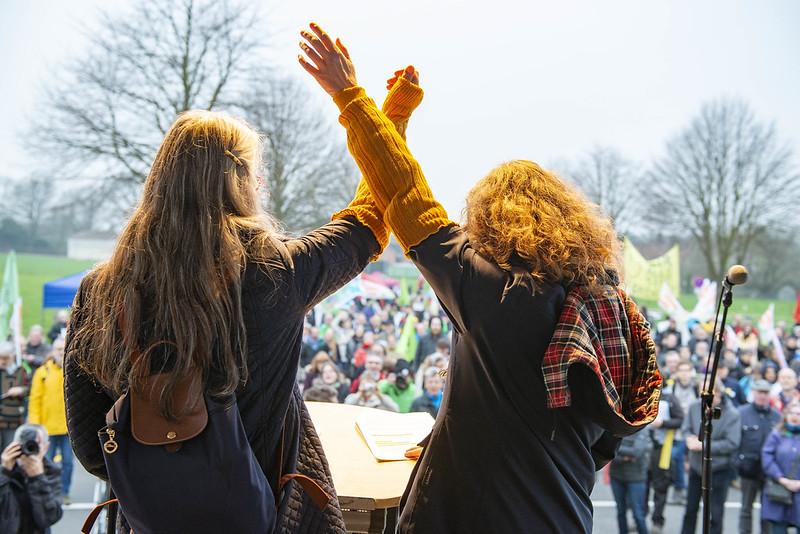 Auf dem Bild sind zwei Personen von hinten zu sehen, die jeweils mit einem Arm zusammen ein gelbes X formen. Sie stehen auf einer erhöhten Bühne, vor der Bühne stehen Menschen in warmer Kleidung. Das Wetter ist etwas trist, im Hintergrund sind unbelaubte Bäume zu sehen.