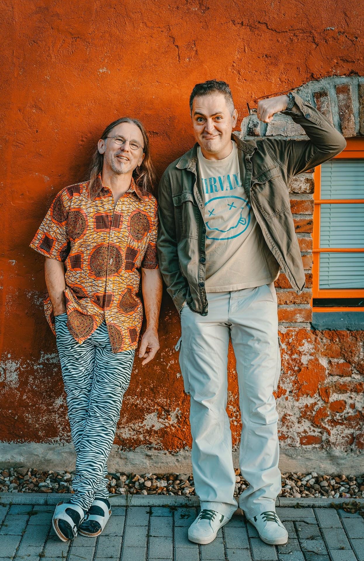 2 Männer mittleren Alters vor einer orangenen Hausfassade, von der teilweise der Putz abbröckelt. Die Person links hat schulterlange Haare, trägt eine Brille sowie ein kurzärmeliges Hemd, eine schwarz-weiß gestreifte Hose, dunkle Socken und hellblaue Birkenstock. Hemd und Hose sind sehr unruhig gemustert. Die Person rechts ist etwas größer, hat kurze dunkle Haare, trägt ein helles T-Shirt und dunkles, geöffnetes Freizeithemd, eine hellblaue Hose und helle Turnschuhe. Das Gesicht ist zu einer leichten Grimasse verzogen, der linke Arm waagerecht angehoben und zum Kopf hin angewinkelt, um zu suggerieren, dass unter dem Hemd Muskeln sind.