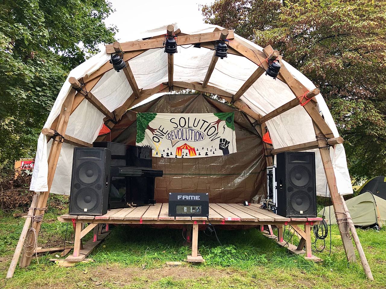 Eine leere, selbst gebaute Bühne aus Holz. Das Dach besteht aus einer weißen Plane, die Rückwand aus brauner Plane. Daran hängt ein Banner mit der Aufschrift "One Solution: Revolution". Auf der Bühne steht ein Klavier sowie mehrere Lautsprecher. Die Bühne ist leicht erhöht, es sind keine Menschen darauf. Sie steht auf einer Wiese, im Hintergrund sind Bäume und ein paar Zelte zu sehen.